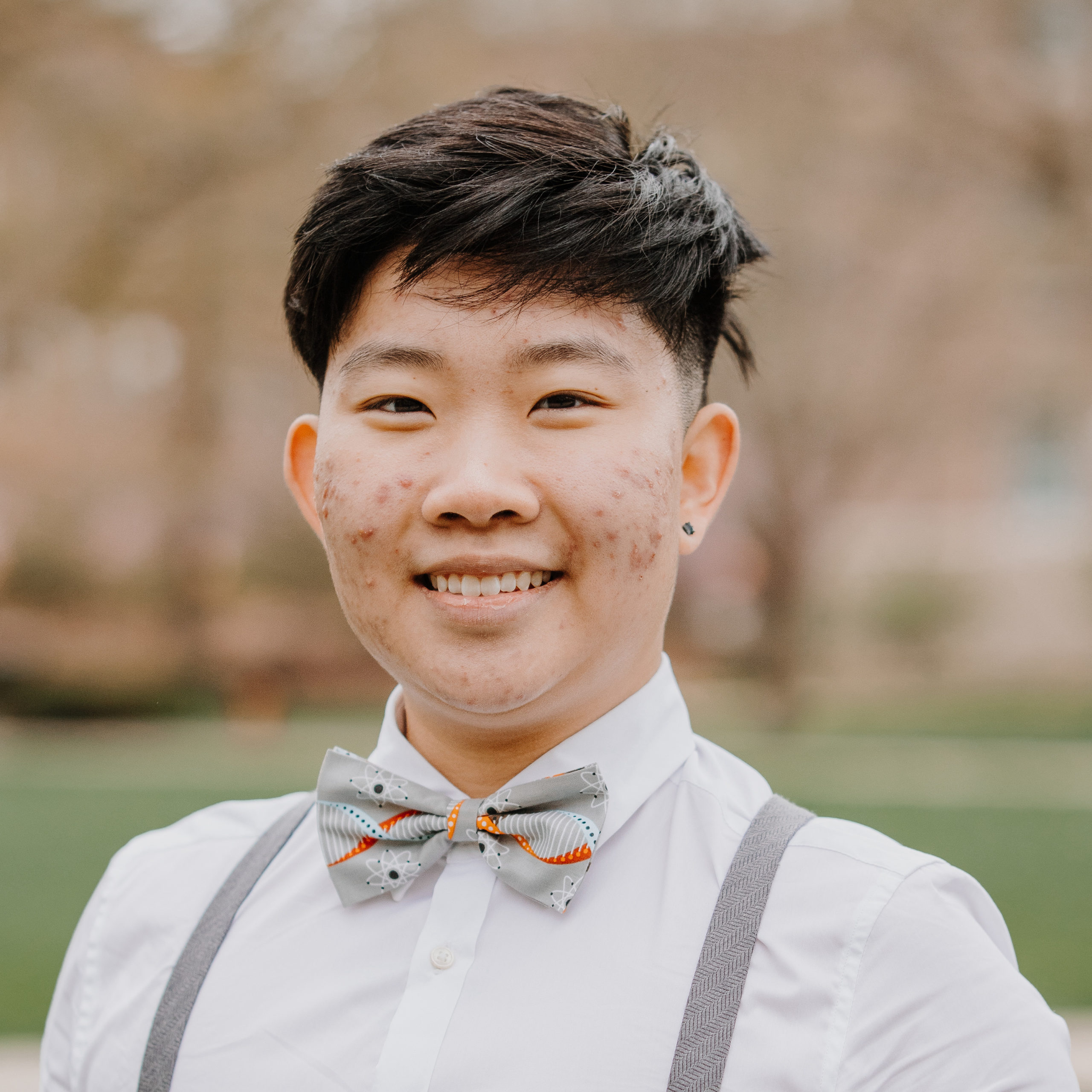 Masculine presenting Asian American person wearing a white dress shirt, a grey bowtie with a blue and orange DNA helix and atom print, and grey suspenders.