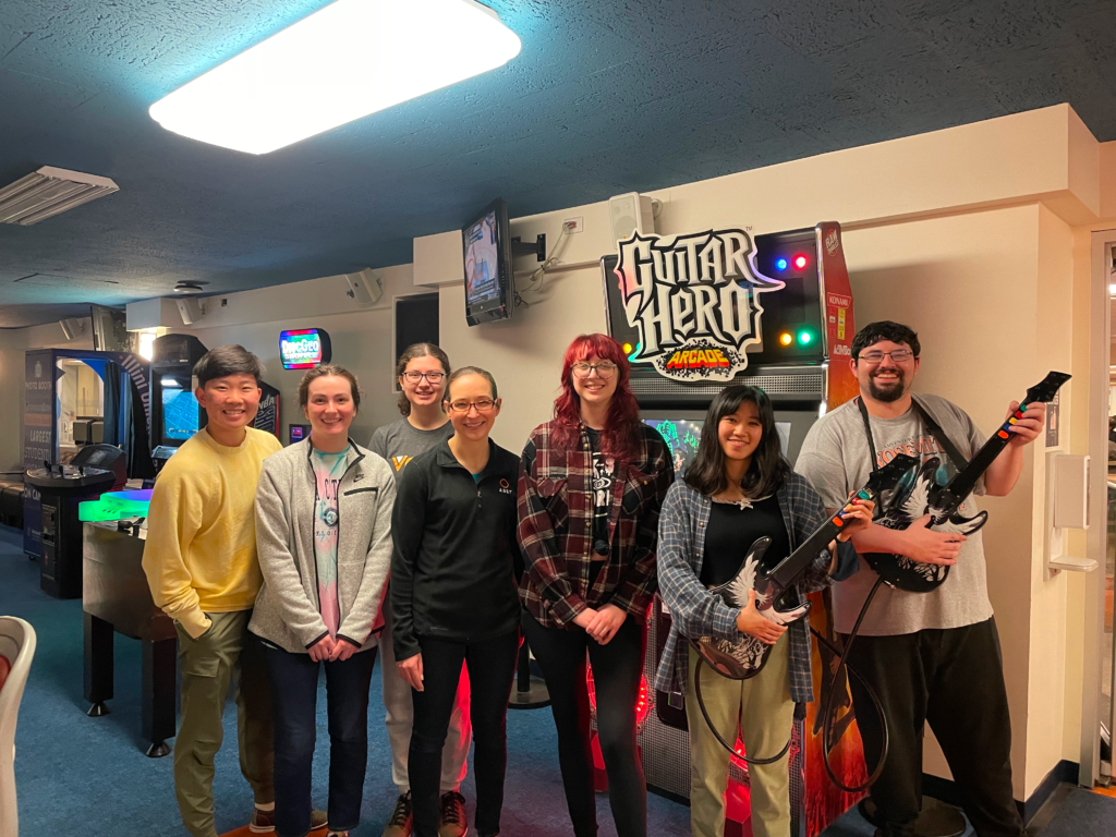 Group photo from 2022 lab bowling party. Evan, Isabel, Sara, Karin, Eileen, Hannah, Joe. Picture is situated inside inside a room with a Guitar Hero sign in the background
