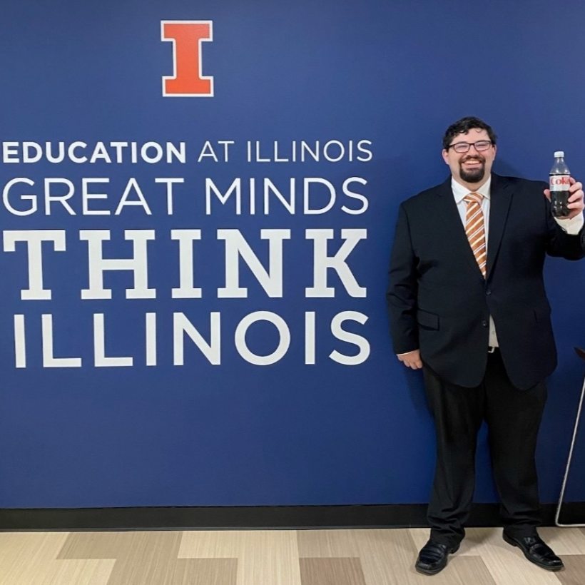KJens lab member Joseph Mirabelli stands in front of a poster that says "Education at Illinois: Great Minds Think Illinois"